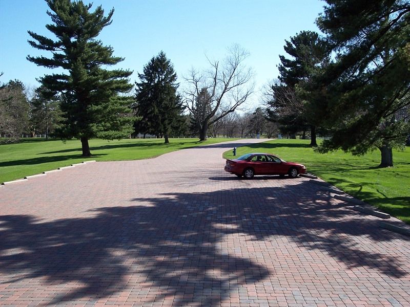 Red car parked on a brick-paved driveway, shadowed by trees on a sunny day.