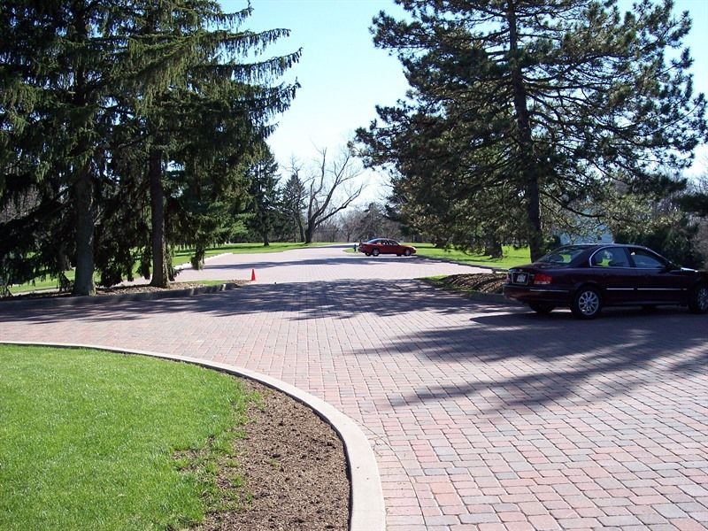 Brick driveway with cars and trees, a grassy lawn on the left and a sunny day.