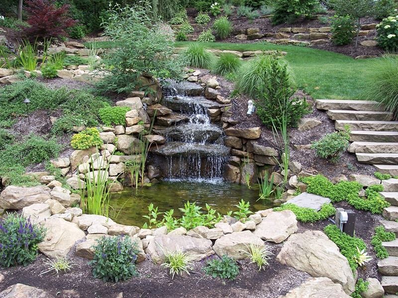 Waterfall cascading into a small pond, surrounded by rocks and lush greenery, with stone steps leading up.