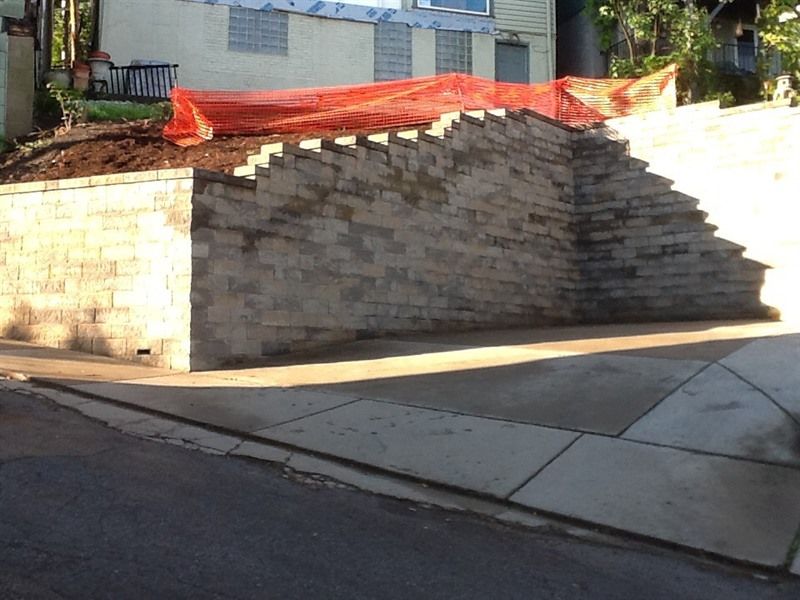 Stone retaining wall with a driveway below and a house in the background. Orange netting is on the top.