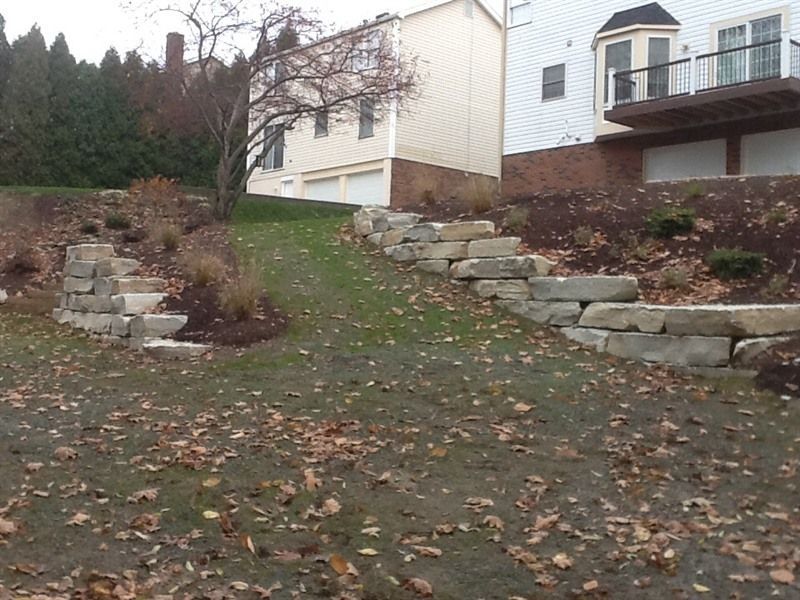 Stone retaining walls and steps on a grassy slope leading to a two-story house.