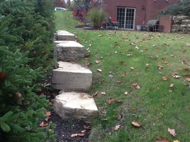 Stone steps ascending a grassy hill next to evergreen shrubs, leading towards a house with a patio.