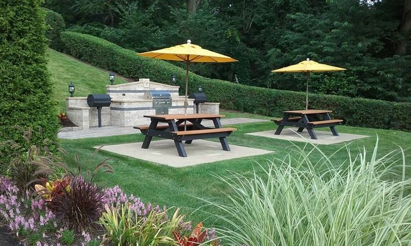 Outdoor picnic area with picnic tables, umbrellas, and grills surrounded by greenery.