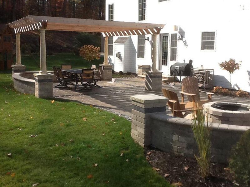 Patio with pergola, seating, fire pit, and retaining walls, adjacent to a white house with green lawn.