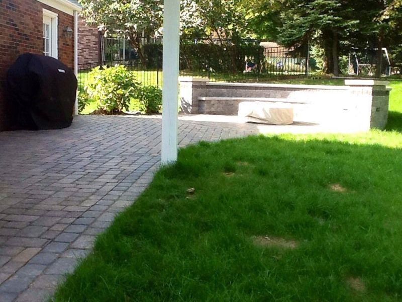 Brick patio with a covered barbecue grill, green lawn, and stone fire pit in a backyard.