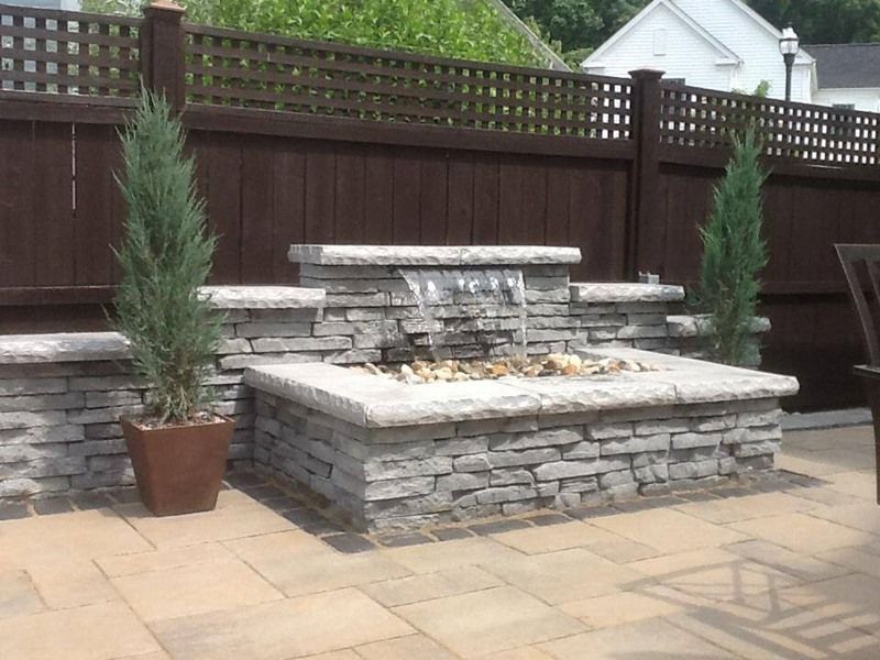 Stone water fountain with a waterfall, two evergreens, and a wooden fence.