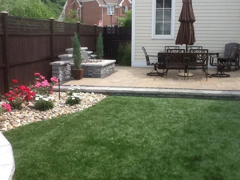 Backyard patio with fire pit, dining set, and manicured lawn. Brown fence, beige patio, and green grass.