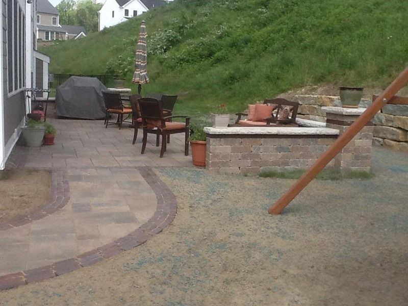 Patio with seating, grill, and fire pit near a grassy hill and house.