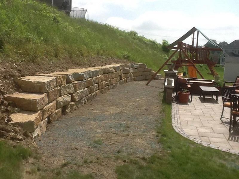 Stone retaining wall next to gravel path, leading to patio with furniture and a playground.