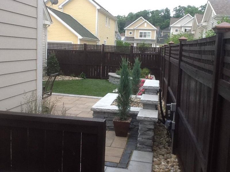 Backyard with brown fence, stone retaining wall, and patio, green grass, and small trees.
