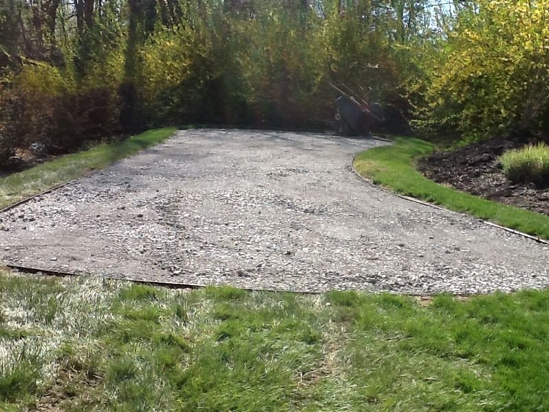 A gravel driveway edged with grass, leading to a wooded area, surrounded by shrubs.