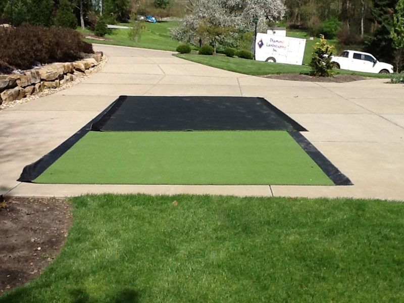 Green and black turf installed in a concrete driveway surrounded by grass and landscaping.