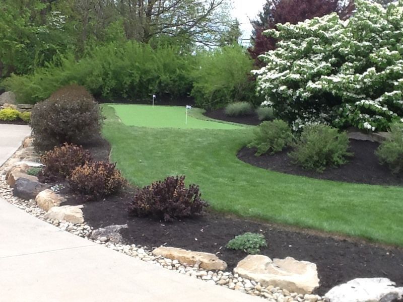 A well-landscaped backyard with a small putting green, surrounded by shrubs, trees, and mulch.