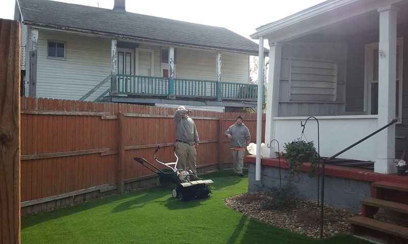 Two people mowing a green lawn in a backyard; a wooden fence and houses are in the background.