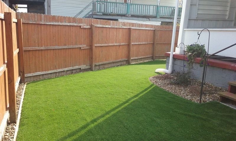 Lush green artificial grass in a small fenced backyard. Brown wooden fence and building in the background.
