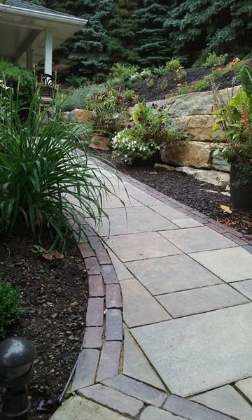 Stone walkway with brick border curving uphill, edged with plants and mulch, beside a hillside.