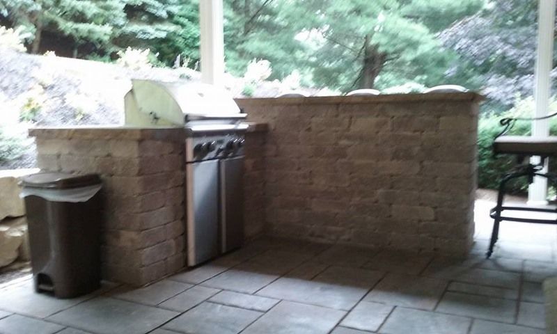 Outdoor built-in stone grilling station with a grill, trash can, and tiled patio.