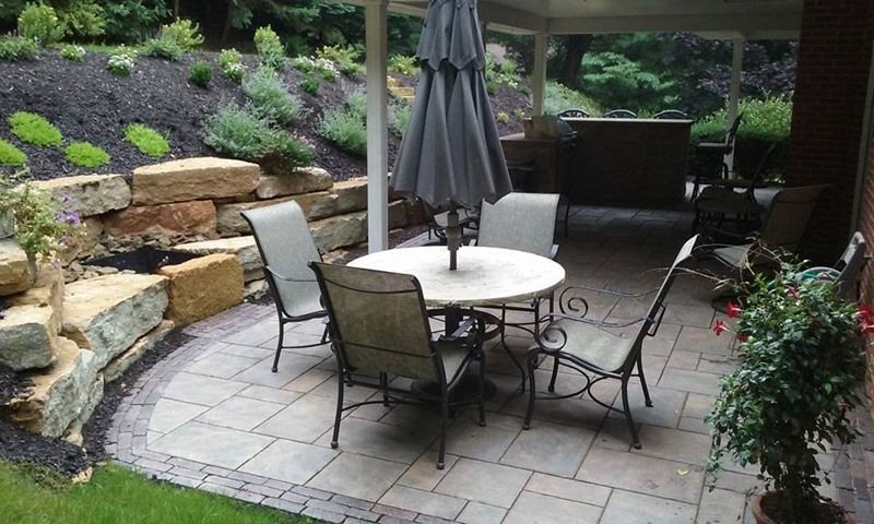 Patio with round table and chairs under a closed umbrella, next to a rock wall and greenery.