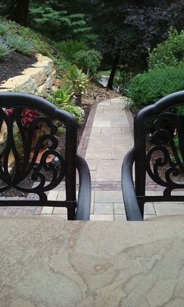 Two ornate black chairs on a stone patio overlooking a brick-lined walkway in a garden.