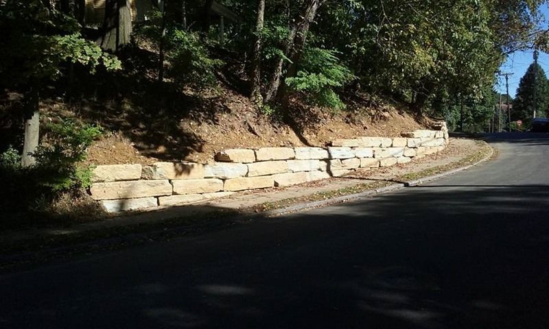 Stone retaining wall along a curved road, with trees and a hillside above.