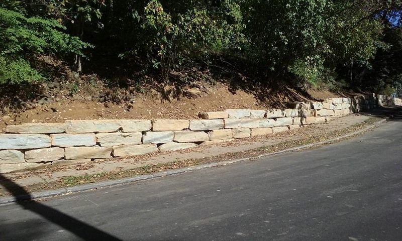 Stone retaining wall along a road, separating the asphalt from a dirt slope with trees.