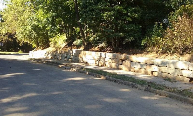 Stone retaining wall along a street, with trees and a sidewalk above.