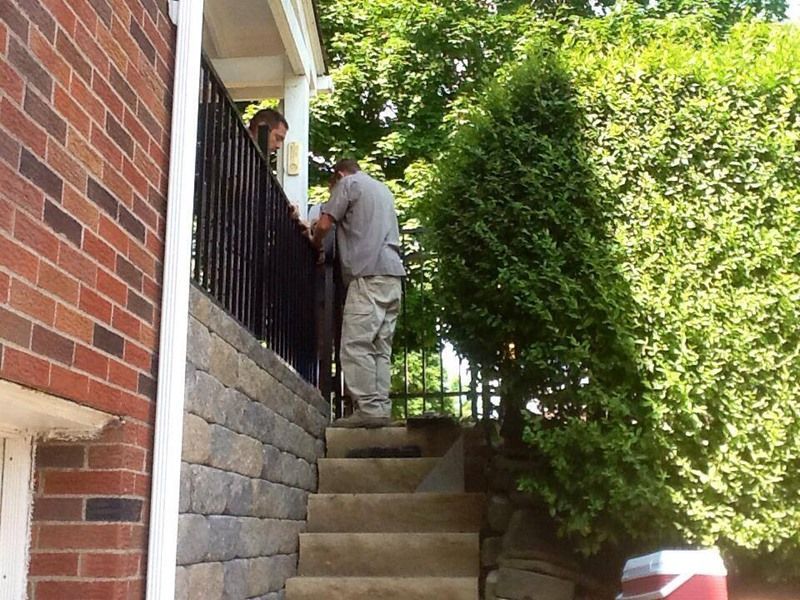 Man on steps talking to person on porch with brick wall and greenery.