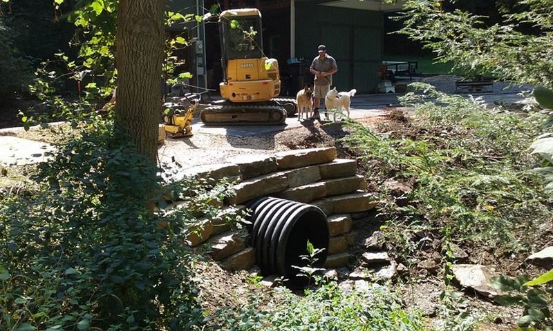 Construction site with a man, two dogs, and a mini excavator. A culvert is installed in a stone retaining wall.