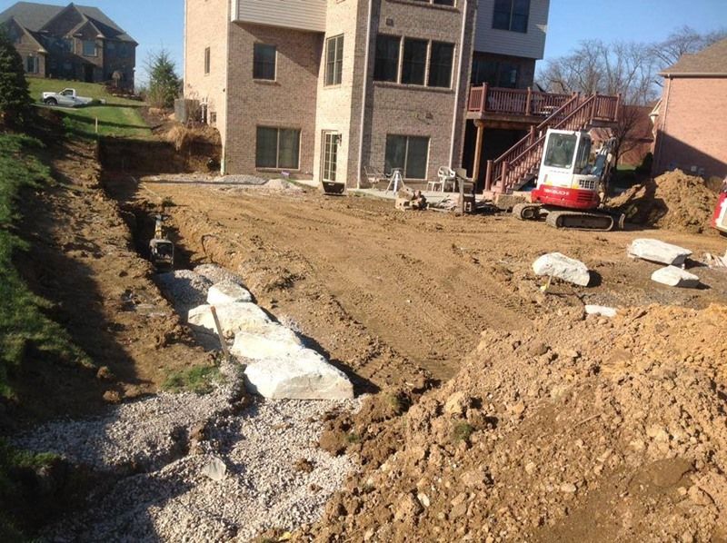 Backyard construction site: excavator and earthmoving near a brick house, retaining wall and gravel.