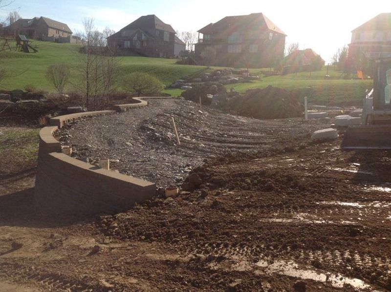 Construction site with retaining wall, gravel, and dirt, with houses in the background on a hill.