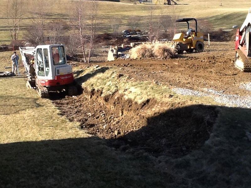 Excavator and roller working on a construction site outdoors, digging trenches in a grassy area.