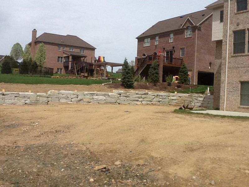 View of two-story brick houses with wooden decks, separated by a dry, graded lot and a stone retaining wall.