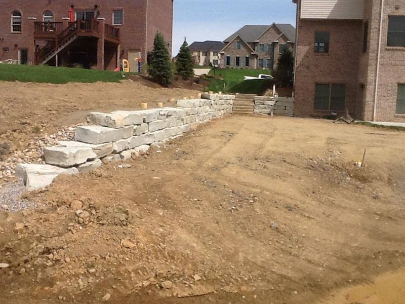 Construction site with stacked stone retaining wall; dirt ground, two-story brick buildings in background.
