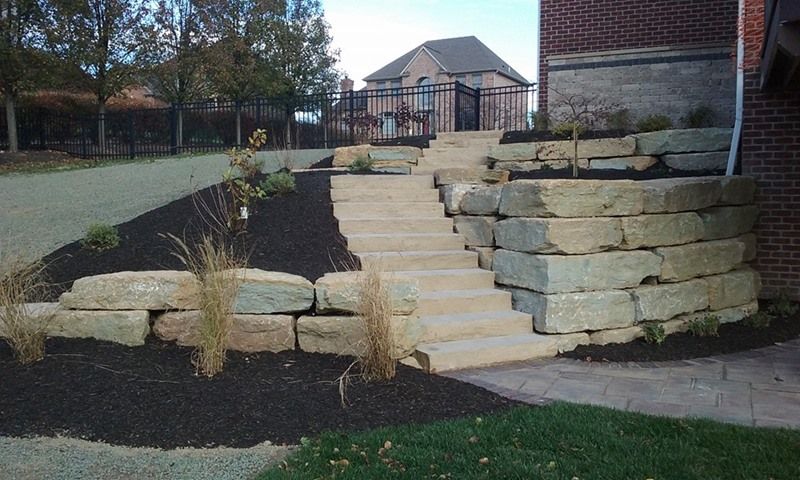 Stone steps leading up, flanked by tiered rock walls with dark mulch and grassy areas.