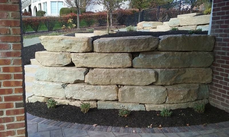 Stone retaining wall in a landscaped area with mulch, plants, and brick building.