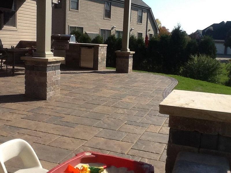 Patio with stone pavers, outdoor kitchen, pillars, and small child's sandbox. Sunny day in suburban backyard.