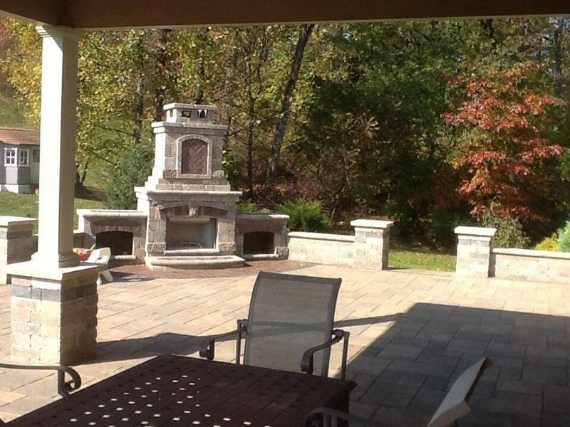 Outdoor patio with stone fireplace, seating area, and surrounding trees. Sunny day.