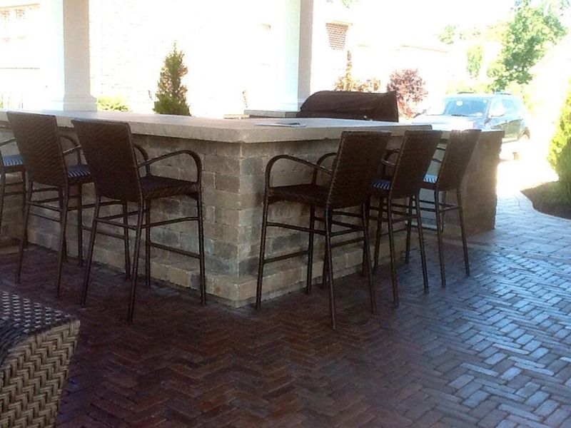 Outdoor bar with seating, built-in grill, and brick patio. Dark chairs line the stone counter.