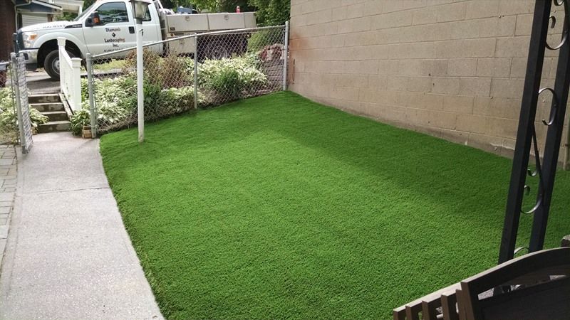 Green artificial turf yard next to a beige wall, a sidewalk, and a white truck.