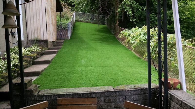 Green artificial turf covers a sloped backyard, bordered by a chain-link fence and stone retaining wall.