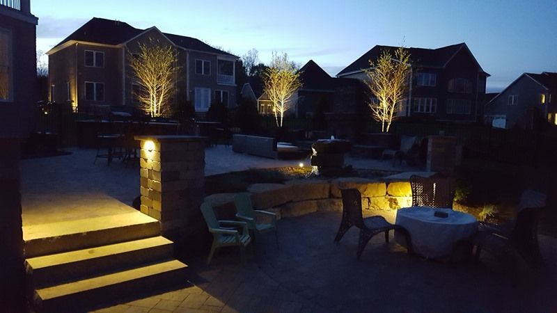 Outdoor patio at dusk, with illuminated trees and buildings in the background. Stairs lead to seating areas, with accent lights.