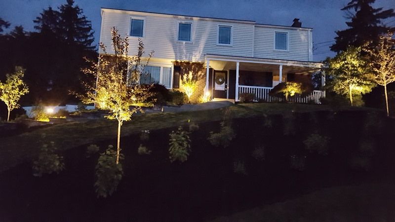 Two-story house illuminated at night by landscape lighting. Trees and bushes in front of the house.