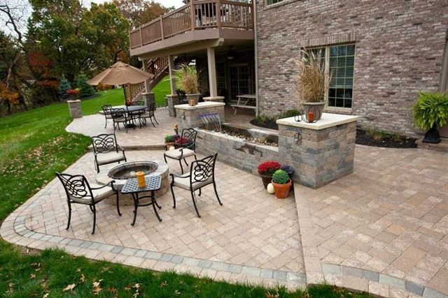 Brick patio with fire pit, seating, and a house in the background. Brown, beige, green, outdoor setting.