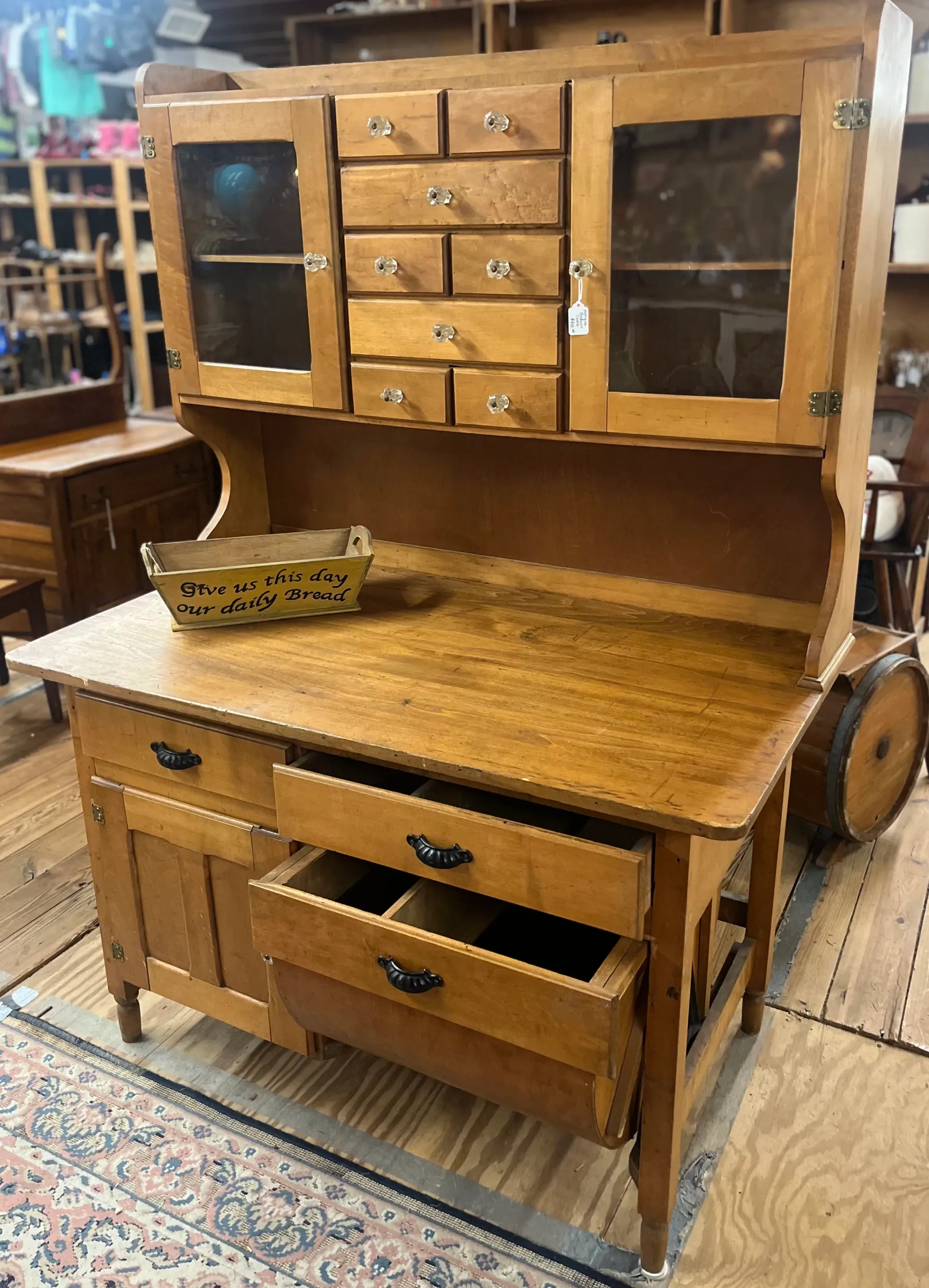 A light oak wooden hutch with glass cabinets, small drawers, and open lower drawers, sitting on a shop floor.