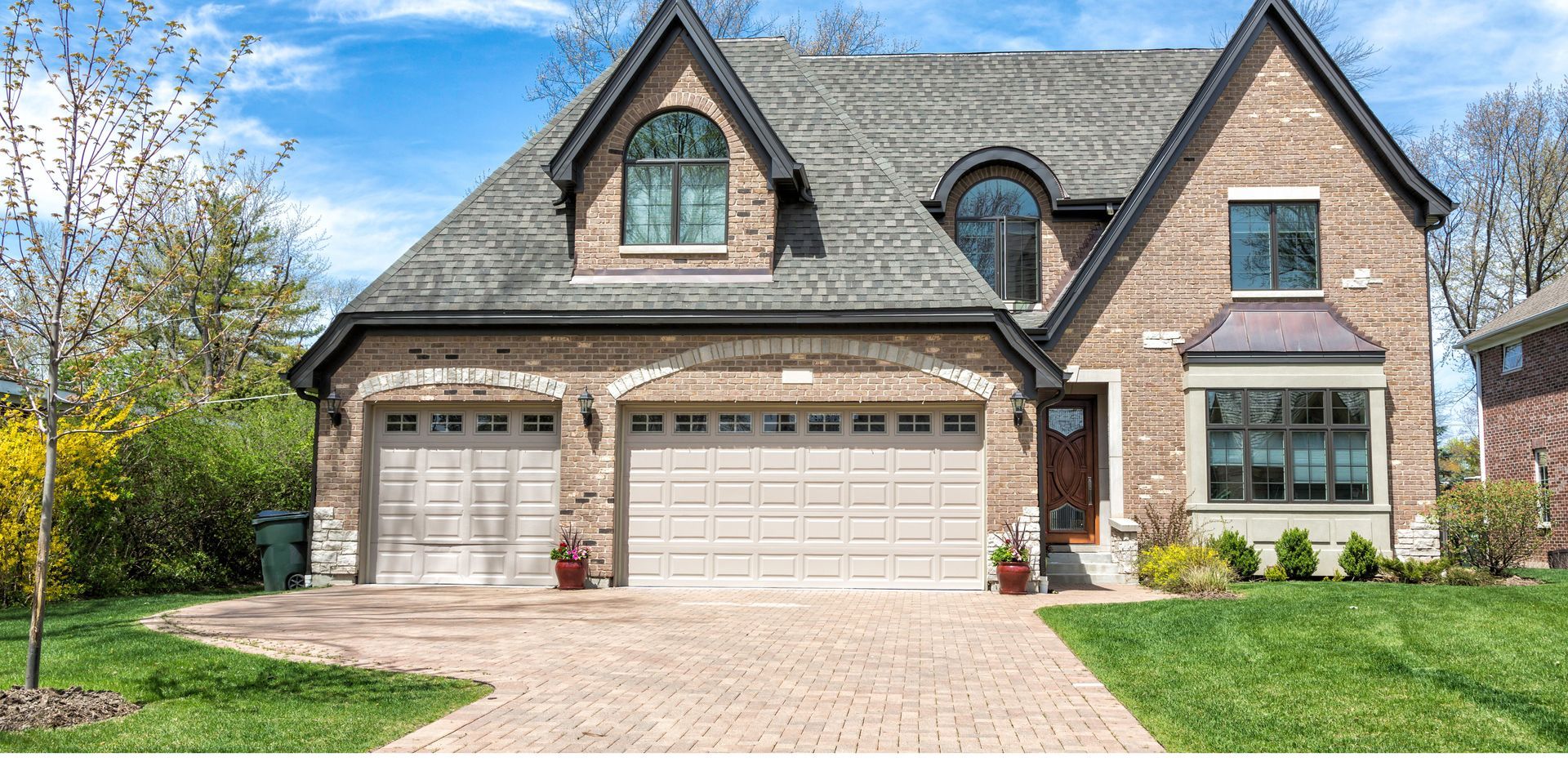 Brick house with two-car garage, brick driveway, and lush green lawn. Blue sky in the background.
