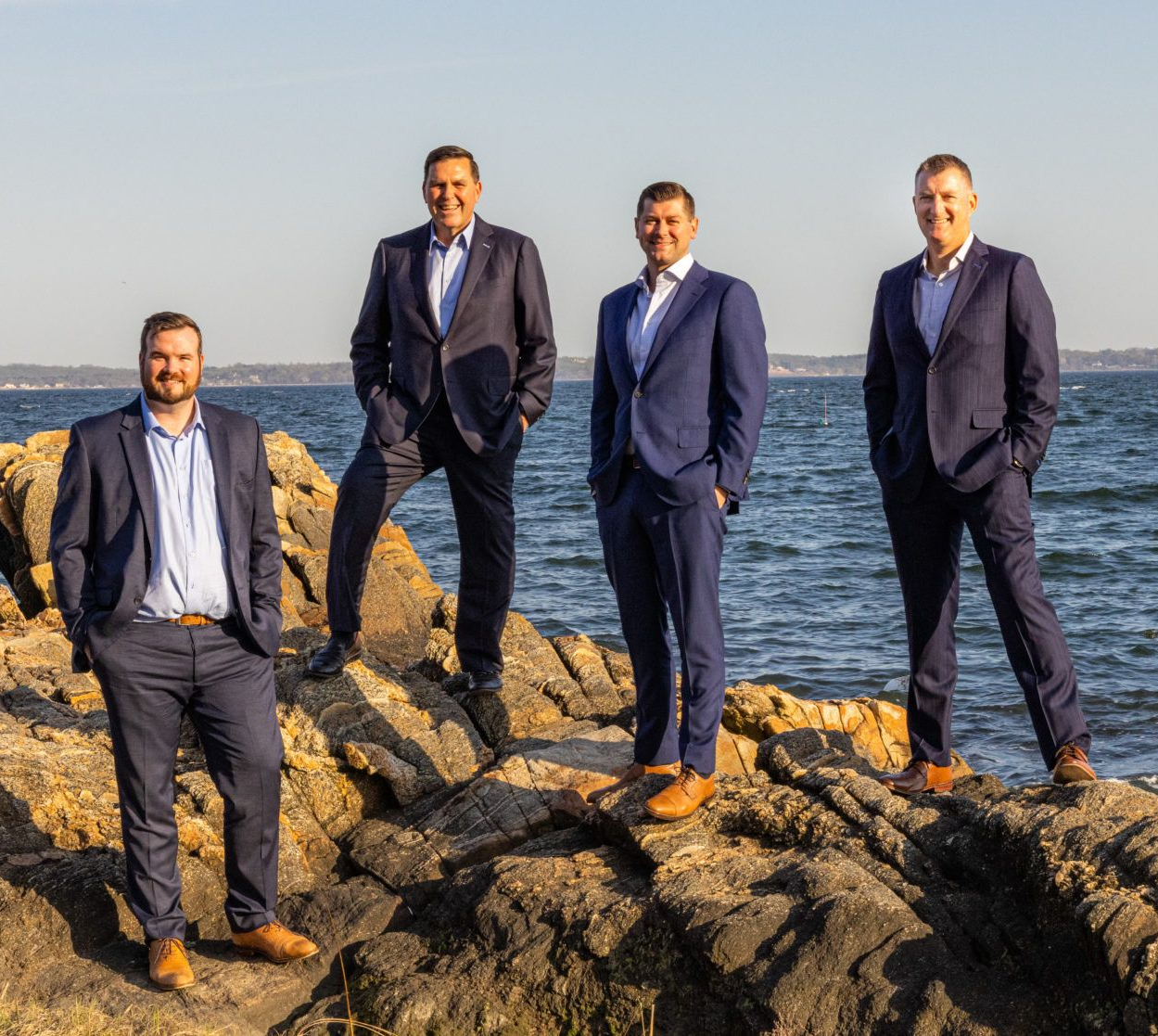 Four men in suits standing on rocks near the ocean, posing for a photo.