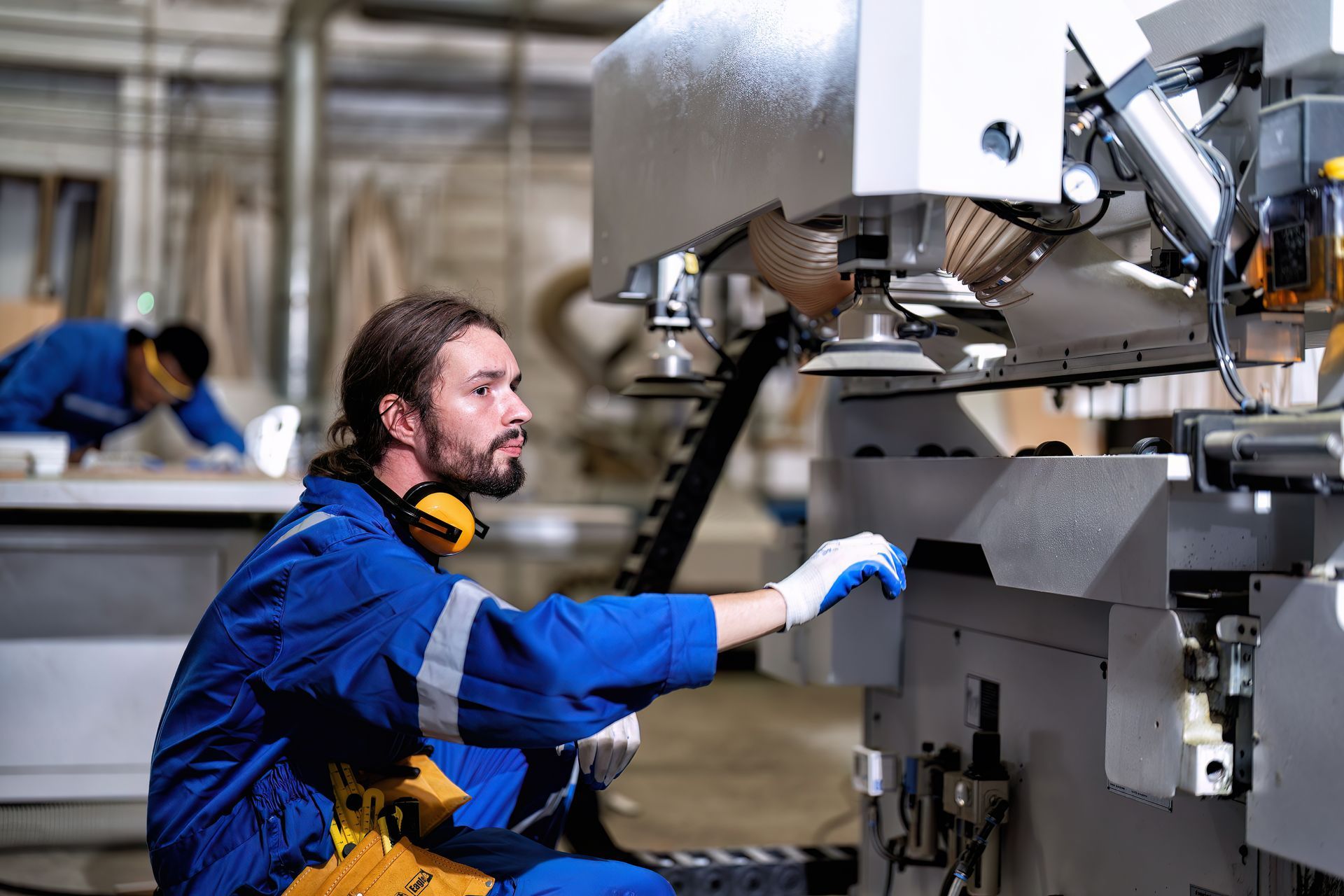 Man in blue overalls operating machinery in a workshop, another worker in background.