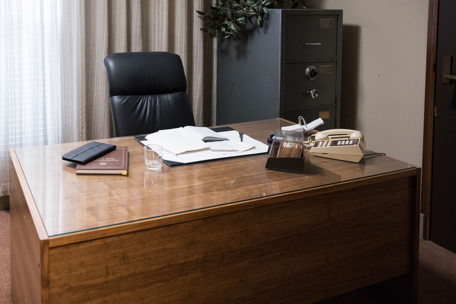 Office desk with papers, phone, and chair; a safe and plant are in the background.