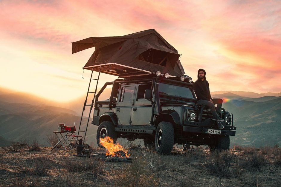 Off-road vehicle with rooftop tent, person leaning on it, campfire, mountains at sunset.