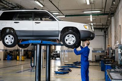 Mechanic in blue overalls working on white SUV lifted on a blue hydraulic lift in a garage.